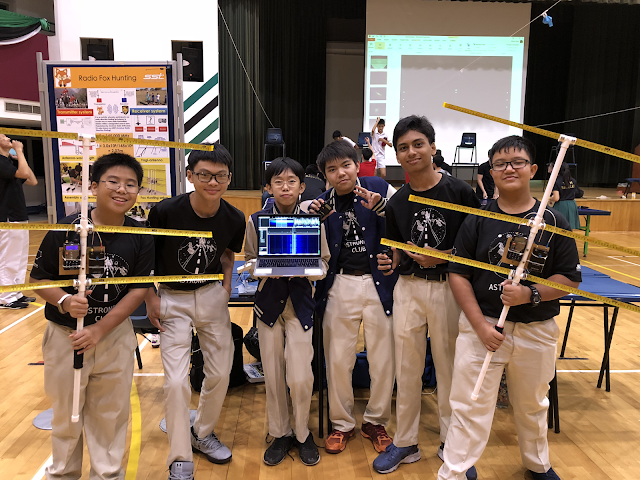 Six boys in matching black club shirts hold antenna rods and a laptop at a Radio Fox Hunting school science fair.